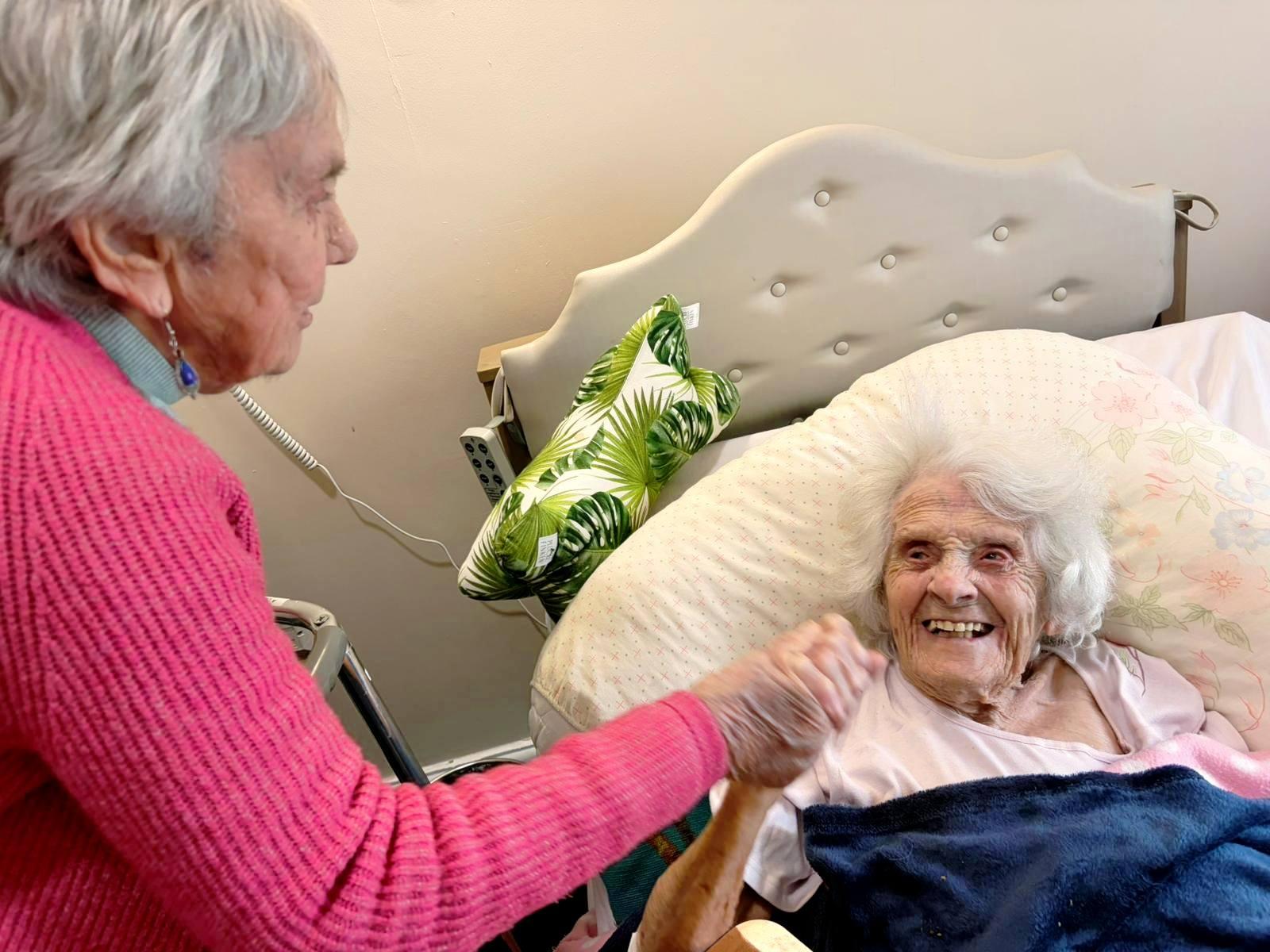 nursing resident in bed holding hands with another elderly lady